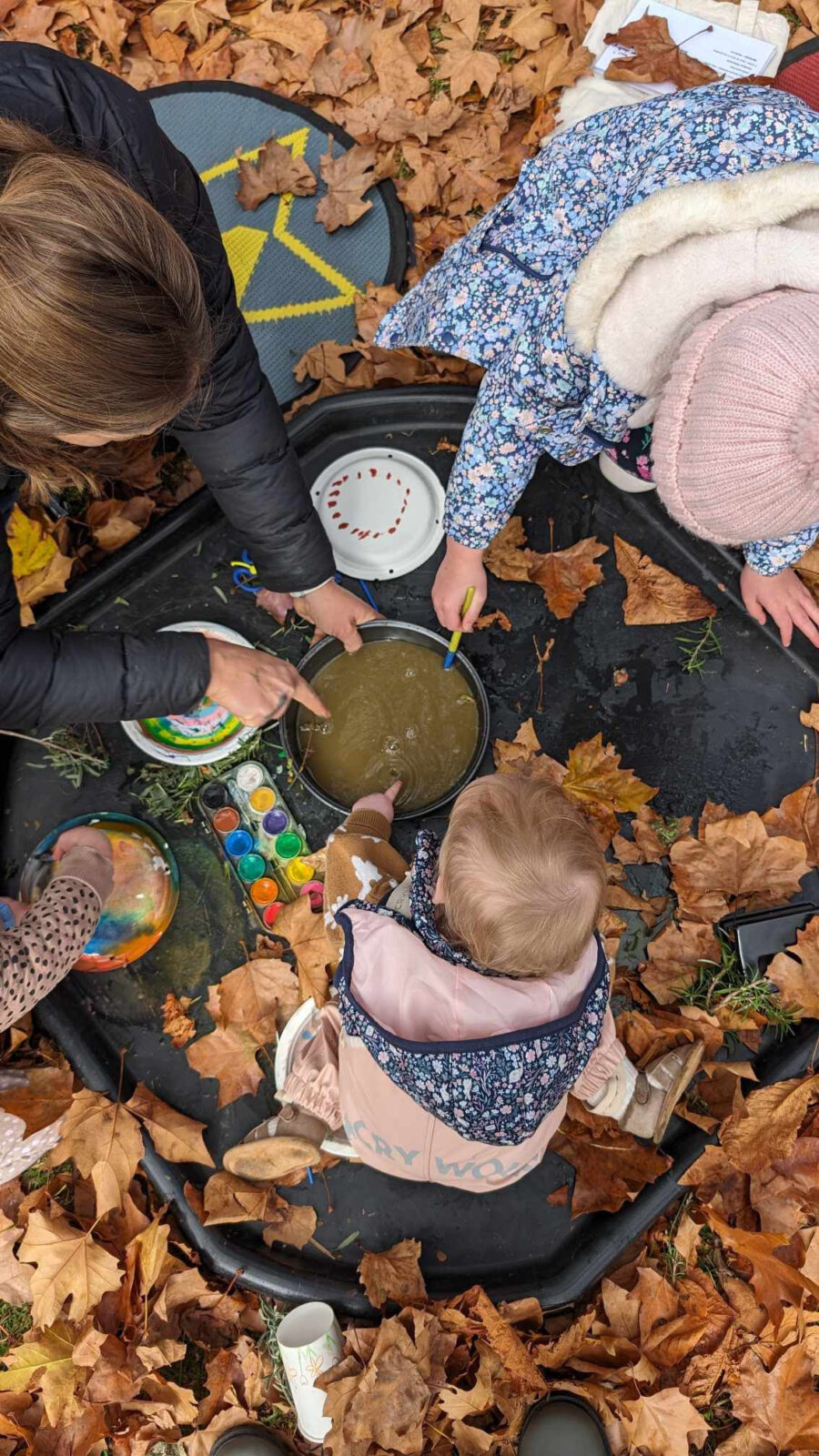 A woman and two children joyfully paint on a leaf-covered table, surrounded by colorful art supplies.