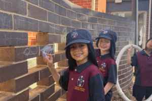 Three children in school uniforms standing by a brick wall, smiling and holding onto it together.