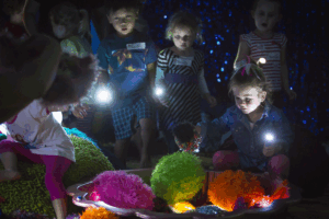 Children joyfully playing with colorful lights in a dimly lit room, creating a magical atmosphere.