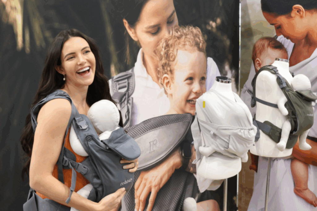 A woman at a trade show holds a baby in a carrier, engaging with the event around her.