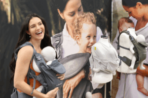 A woman at a trade show holds a baby in a carrier, engaging with the event around her.