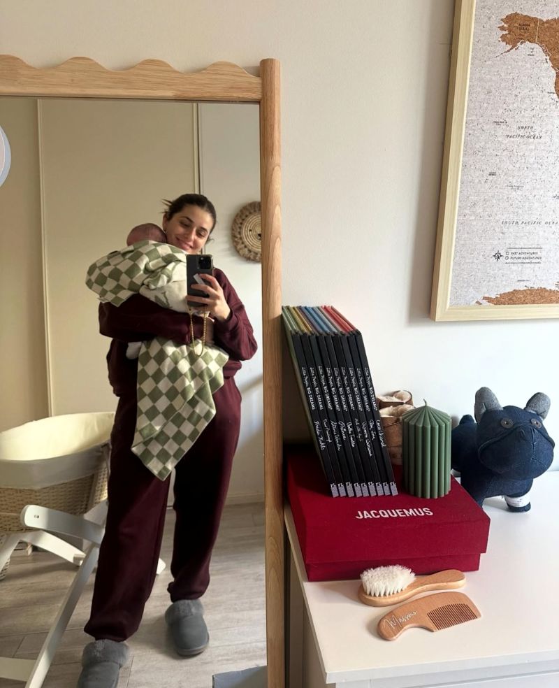 A woman stands in front of a mirror, holding a baby while looking at their reflection together.