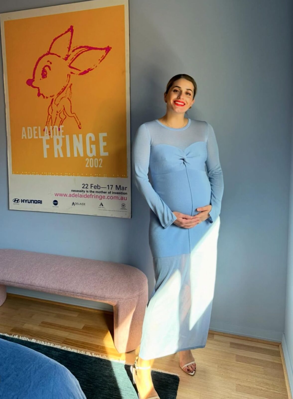 A pregnant woman in a blue dress stands beside a colorful poster, smiling and looking at the camera.