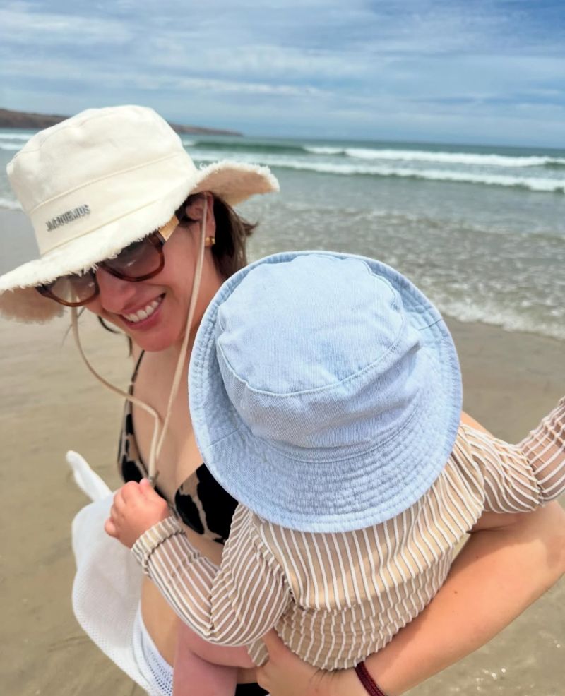 A woman in a hat and sunglasses holds a baby while enjoying a sunny day at the beach.