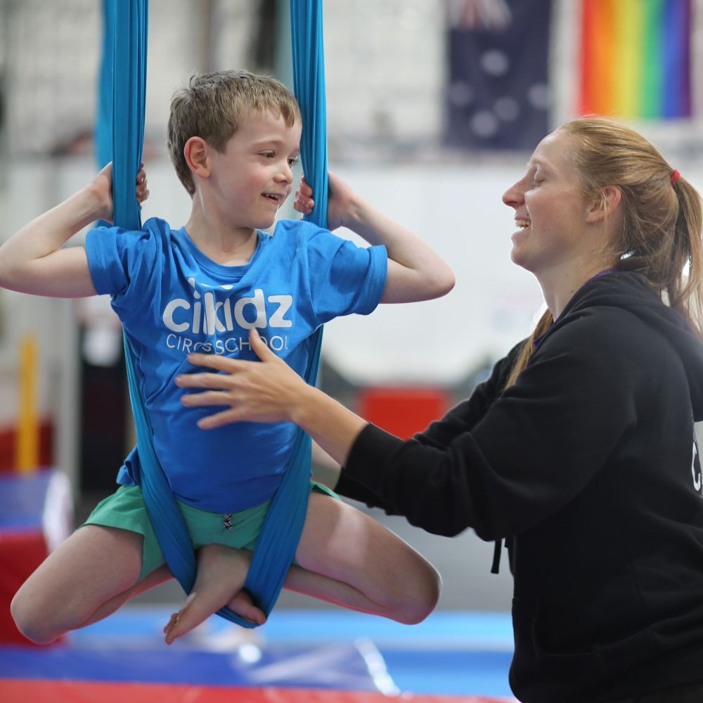 A woman assists a young boy as he jumps on a trampoline, both smiling and enjoying their time together.