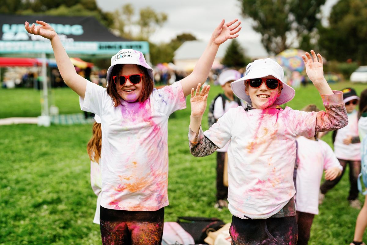 Two young children in white shirts and hats, smiling with colorful paint splatters on their clothing.