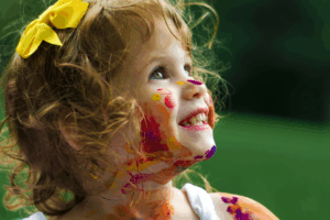 A little girl with colorful paint smeared on her face, smiling joyfully at the camera.