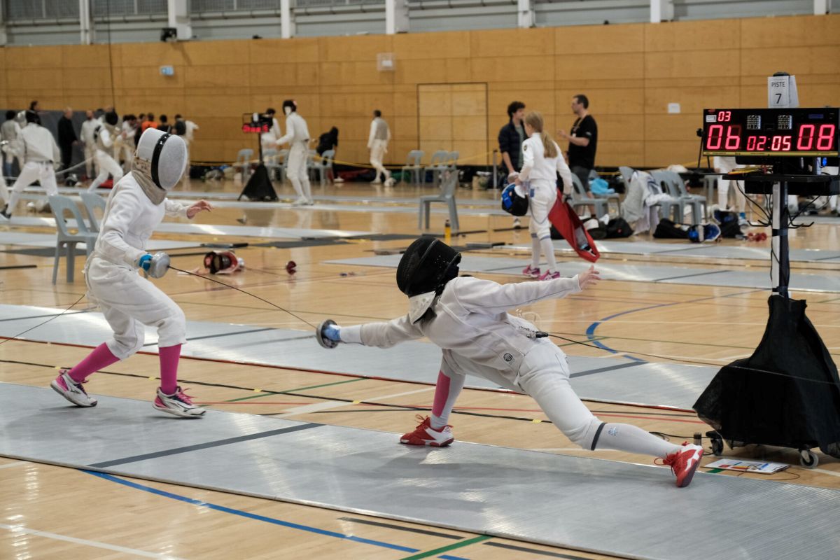 Two fencers in full gear sparring on a fencing court, focused and engaged in their match.
