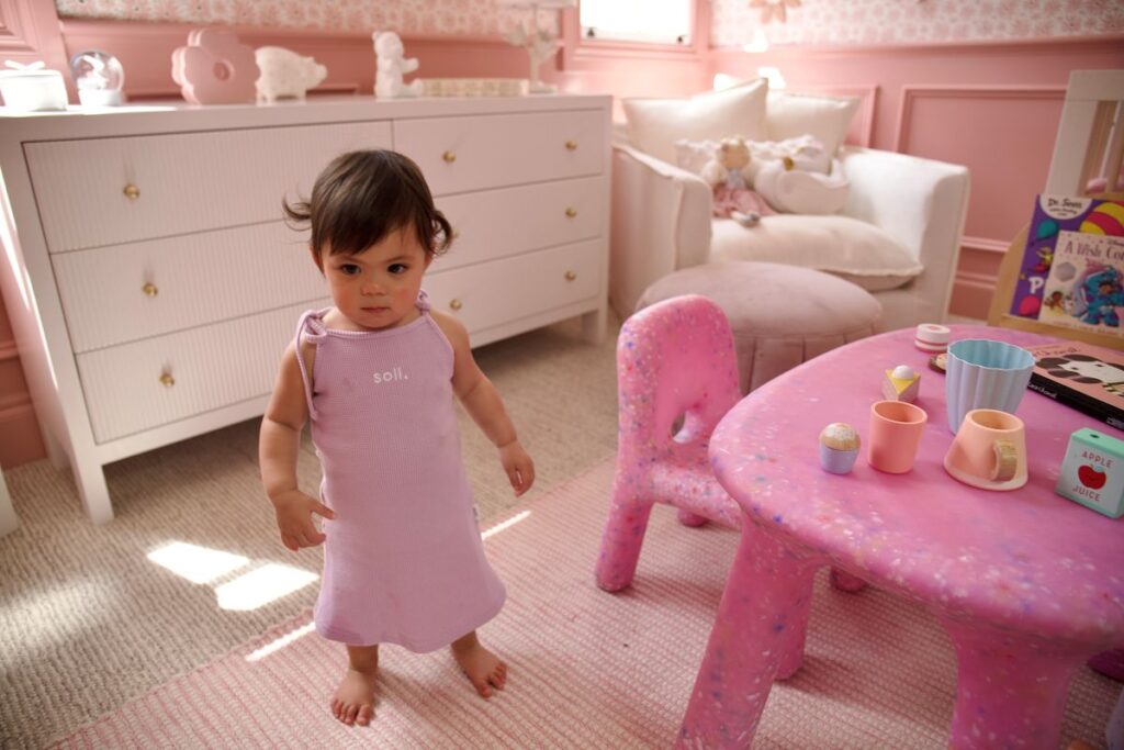 A baby girl in a pink dress stands in front of a matching pink table, smiling and looking at the camera.