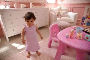 A baby girl in a pink dress stands in front of a matching pink table, smiling and looking at the camera.