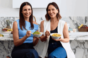 Two women sit in a kitchen, smiling and holding bowls of food, enjoying a moment together.