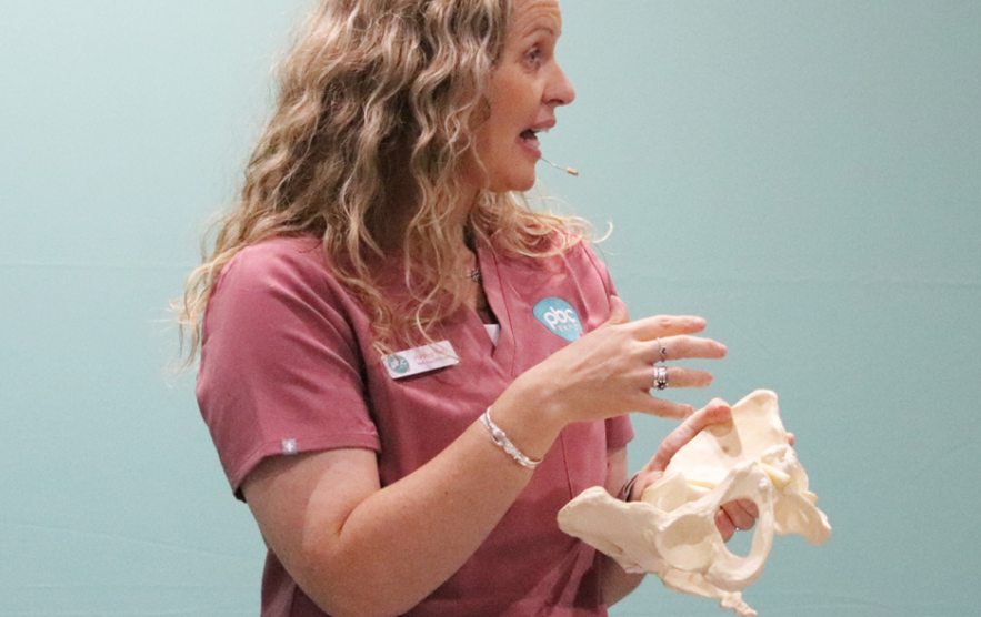 A woman in a pink shirt holds a human skeleton model, smiling as she demonstrates its features.