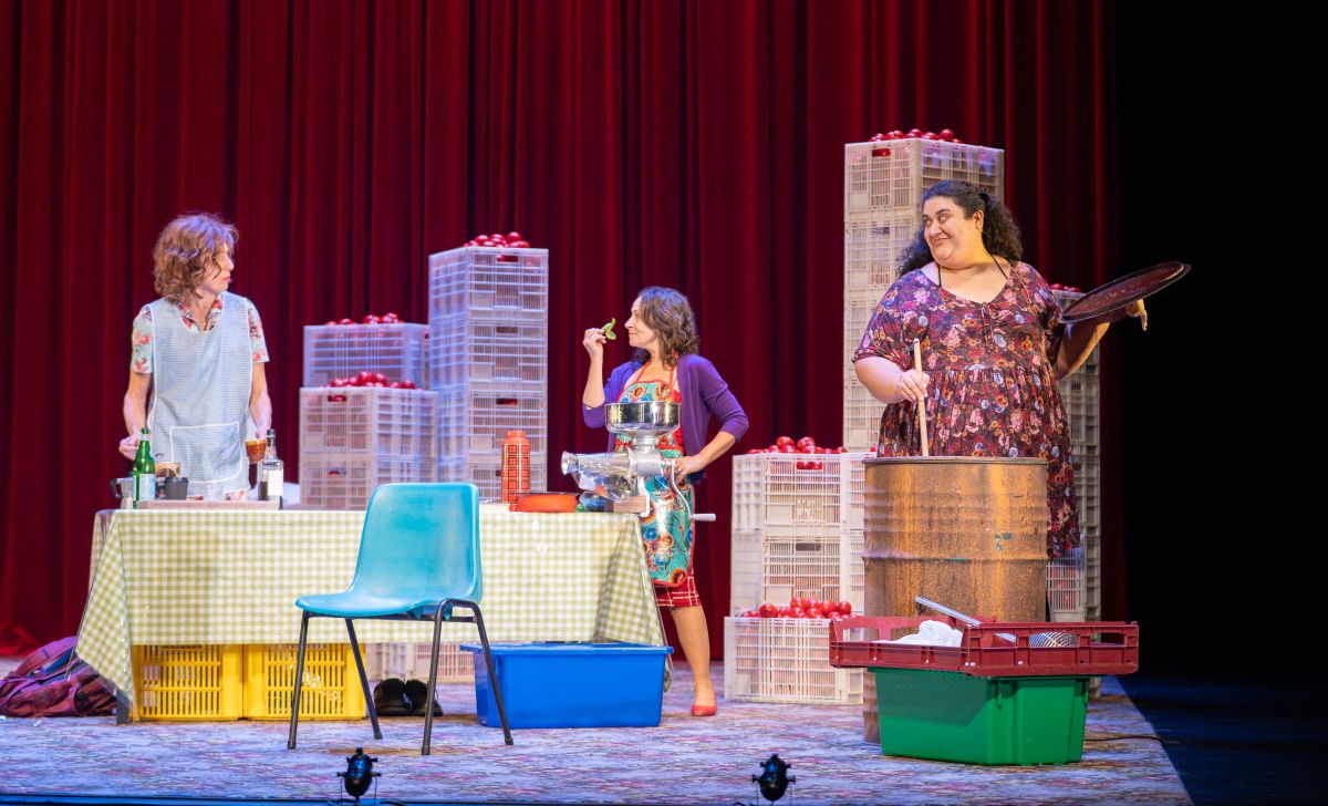 Three women gather around a table filled with boxes, while one woman in a dress stands prominently among them.