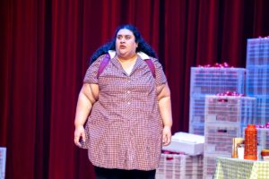 A woman in a dress stands on stage beside several stacked boxes, engaging with the audience.