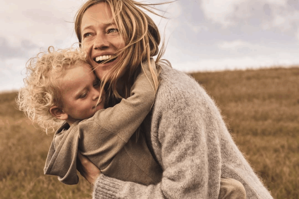 A woman holds a child in her arms while standing in a lush green field under a clear blue sky.