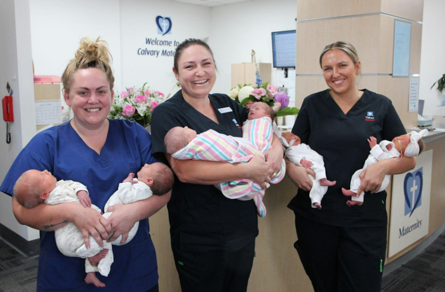 Three women in scrubs smiling while holding newborns in their arms, showcasing a caring and nurturing environment.