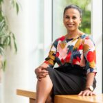 A woman sitting on a bench, gazing out a large window with natural light streaming in.
