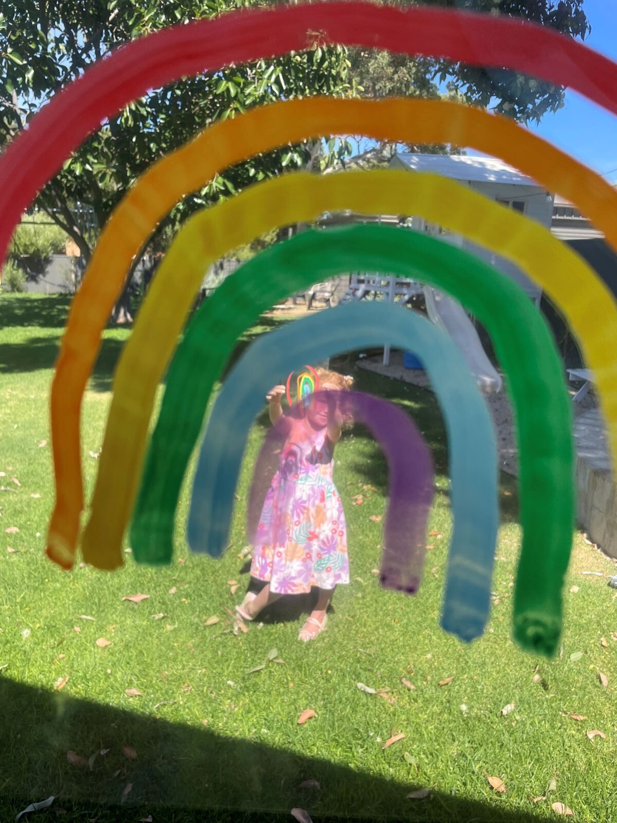 A child stands joyfully in front of a colorful plastic rainbow, smiling and reaching out towards it.