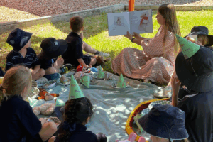 A woman reading a story to children gathered around her on a colorful blanket in a sunny outdoor setting.