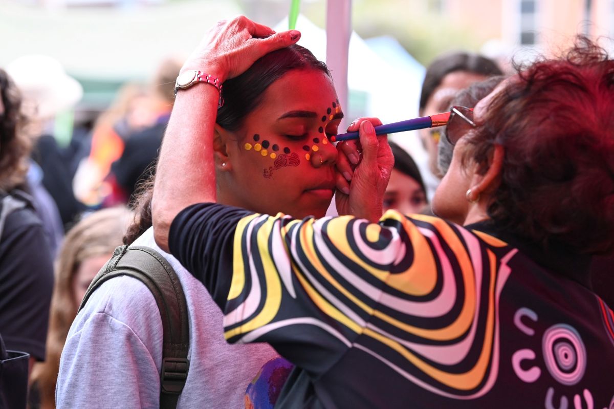 A woman smiles as an artist paints her face at a lively outdoor event.