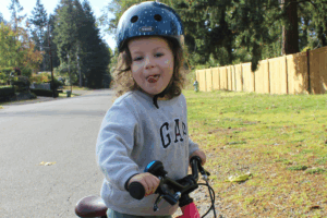 A little girl joyfully riding a bike on a smooth paved road under a clear blue sky.