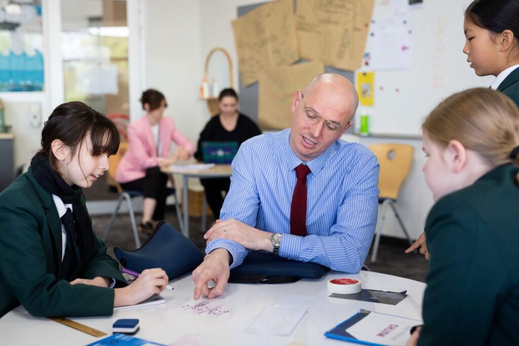 A man in a suit and tie sits at a table, engaging in conversation with a group of students.