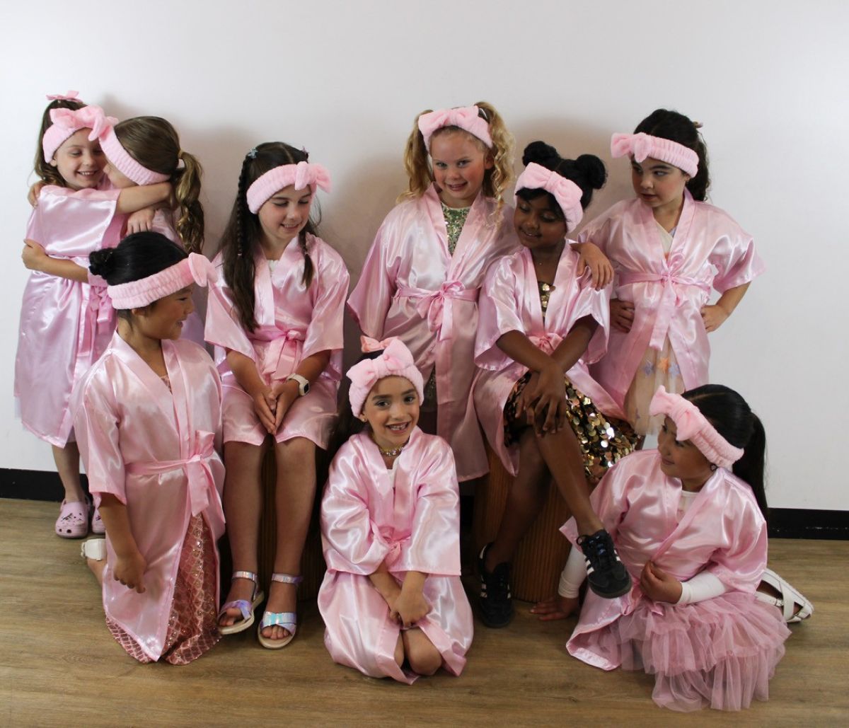 A group of young girls wearing matching pink robes, smiling and standing together in a cheerful setting.