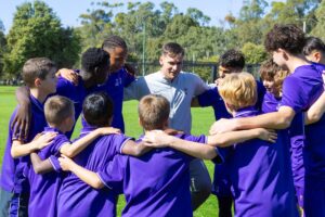 A group of young boys in purple shirts huddle closely together, smiling and engaging with one another.