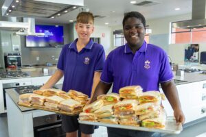 Two boys in purple shirts proudly hold trays filled with sandwiches.