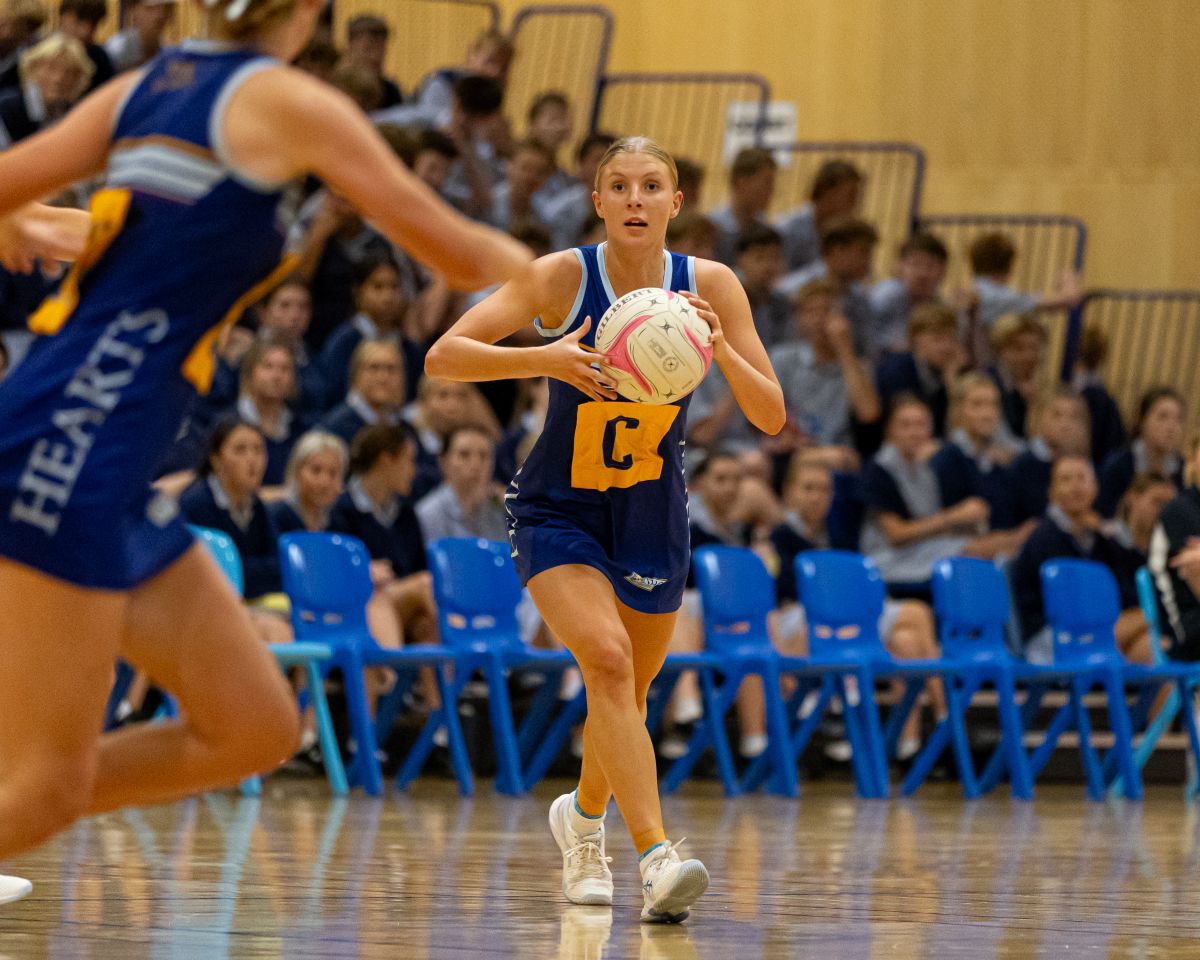 A female netball player in a blue and yellow uniform runs with the ball, focused on the game ahead.