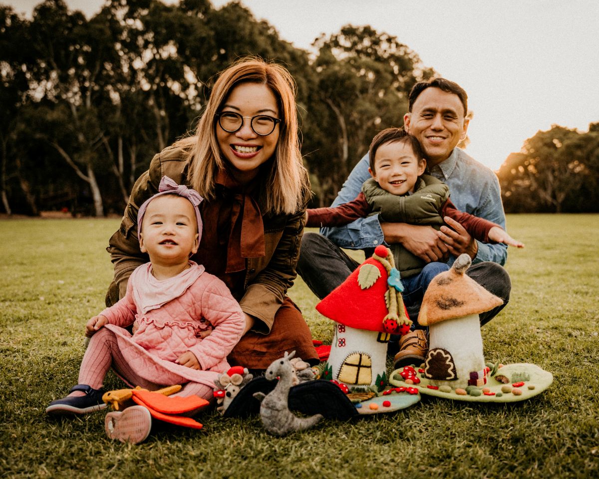 A family smiles together for a photo in a sunny park, surrounded by green grass and trees.