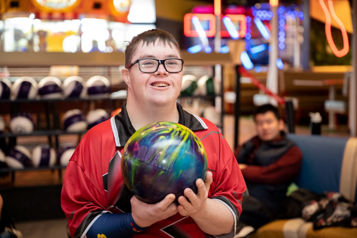 A young man with glasses is holding a bowling ball, preparing to bowl in a bowling alley.