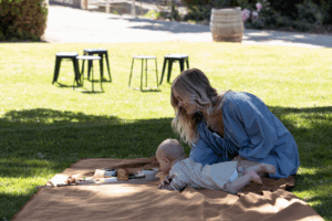 A woman sits on a picnic blanket with a baby, enjoying a sunny day outdoors.