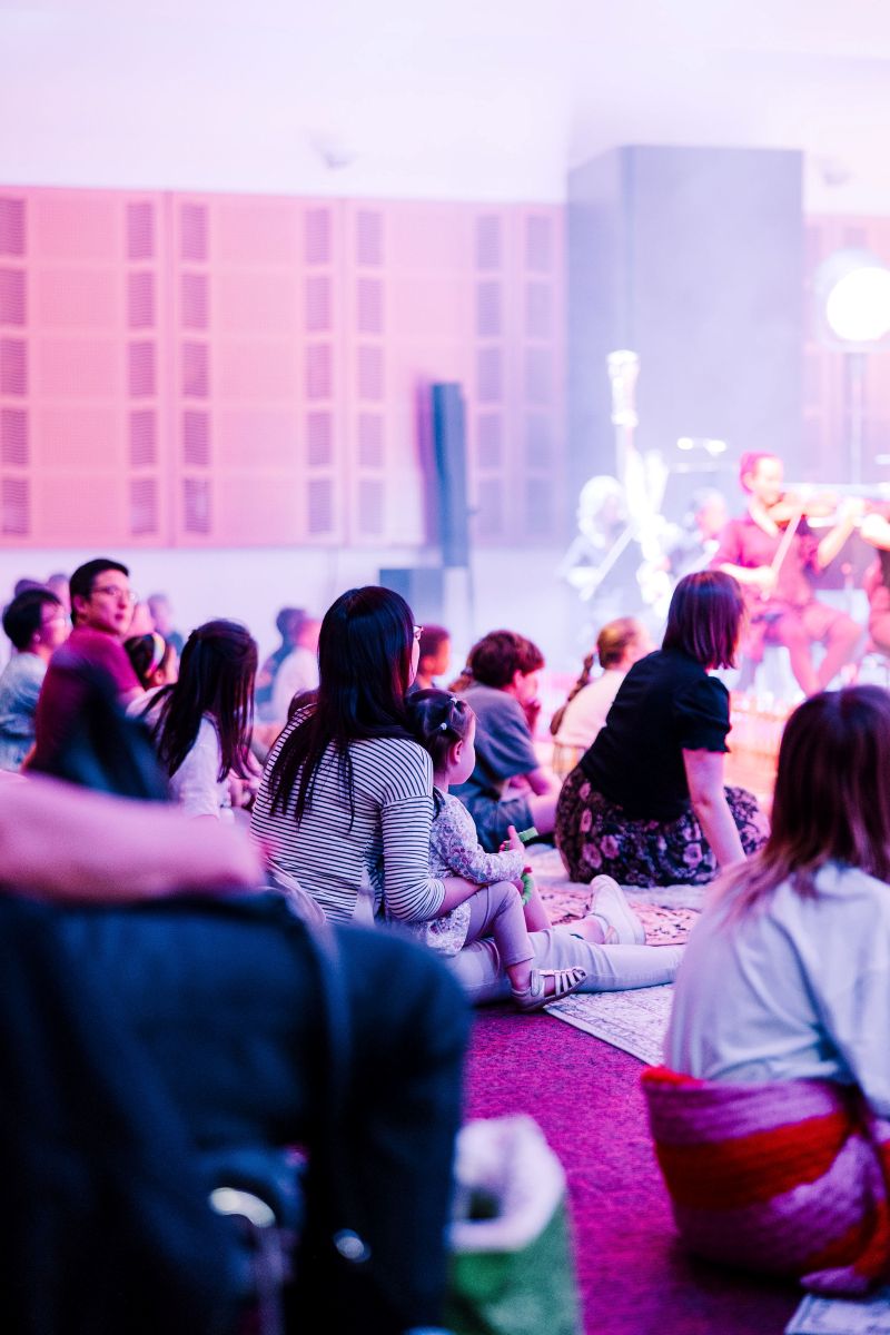 A group of people sitting on the floor in a large auditorium, listening to music.