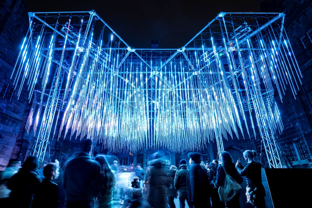 People stroll through a city beneath a large illuminated structure, creating a vibrant urban atmosphere at night.