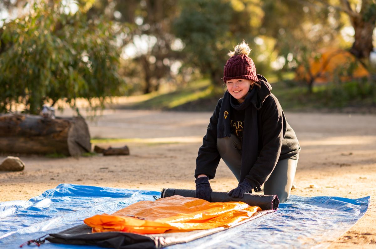 A woman kneels on the ground beside an orange sleeping bag, preparing for a camping trip in a natural setting.