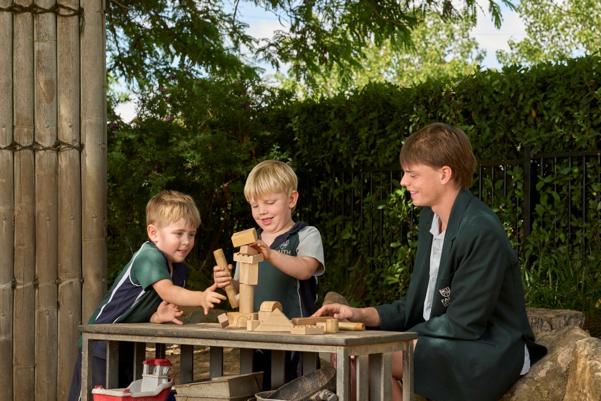 Two children happily playing with colorful wooden blocks on a table, building structures together with a senior student looking on.