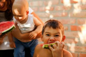 A woman holds a child while enjoying a slice of watermelon on a sunny day.