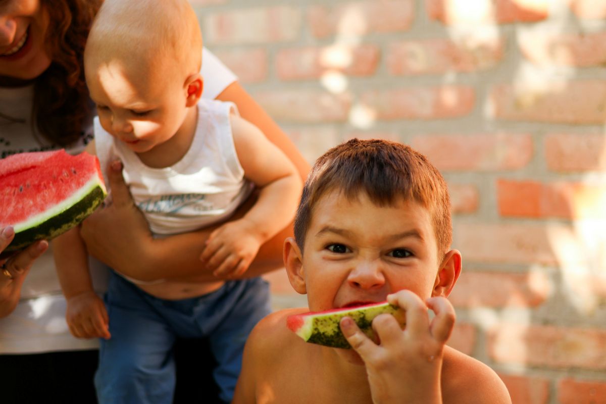 A woman holds a child while enjoying a slice of watermelon on a sunny day.