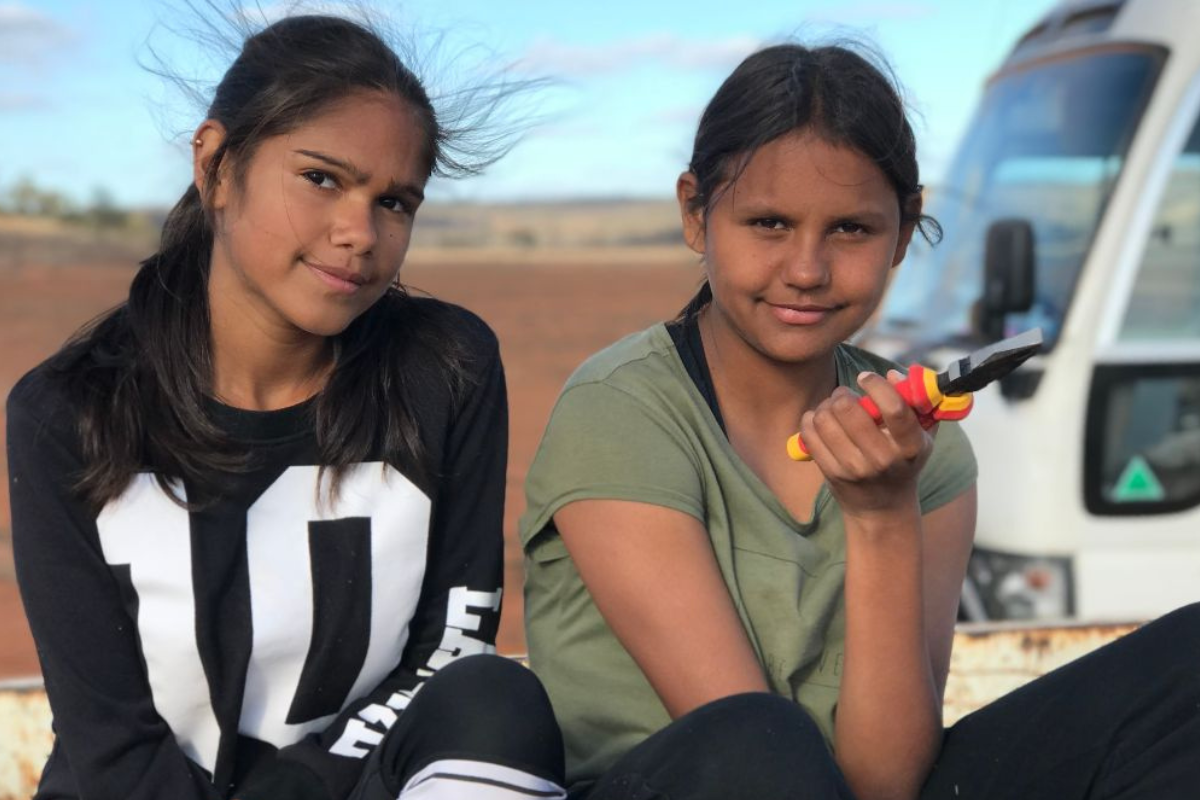 Two young girls sitting on the back of a truck, smiling and enjoying a sunny day together.