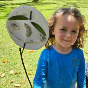 A young girl smiles while holding a paper plate featuring a colorful bird illustration.