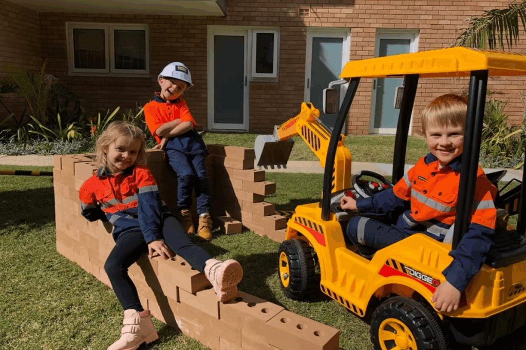 Three children happily sitting on a bright toy construction vehicle, smiling and enjoying their playtime outdoors.