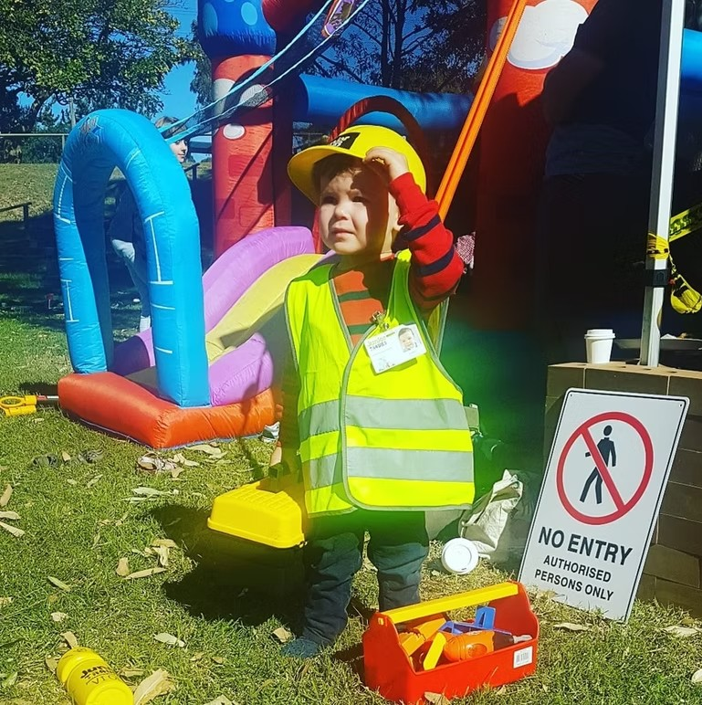 A young boy in a safety vest stands beside a colorful bouncy castle, smiling and ready to play.