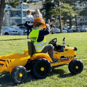 A young boy happily riding a toy tractor in a sunny park, surrounded by green grass and trees.