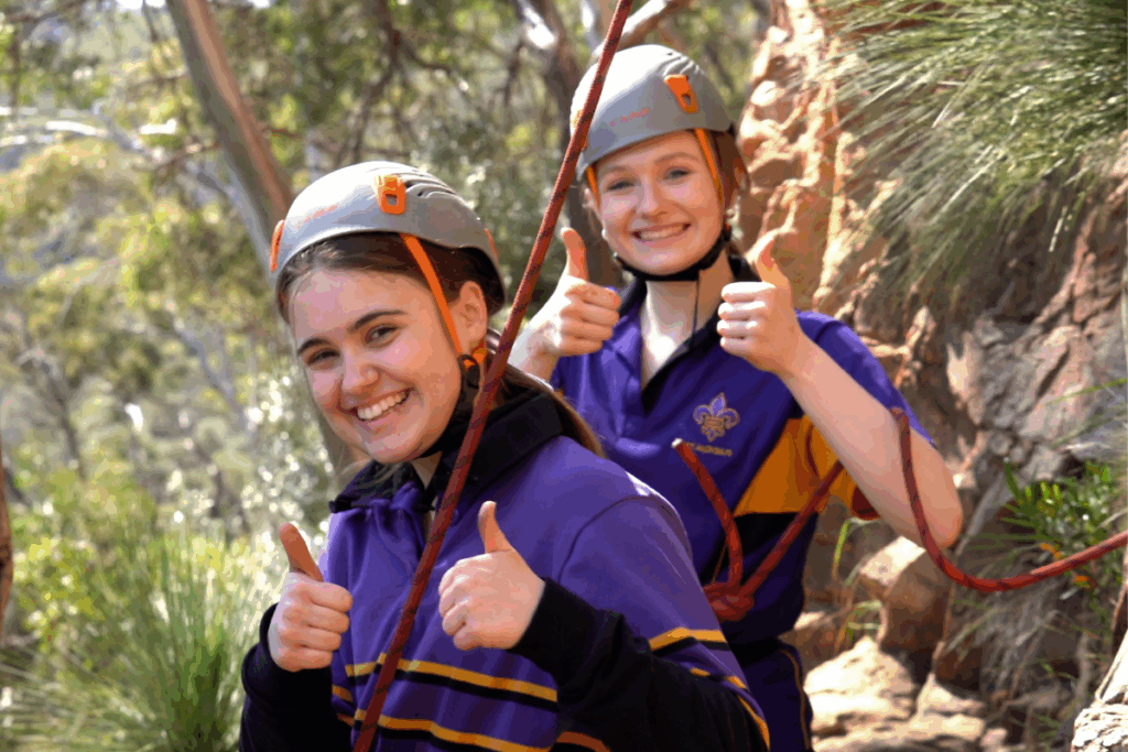 Two young women in purple shirts and helmets navigate a rope course, focused and enjoying the outdoor adventure.