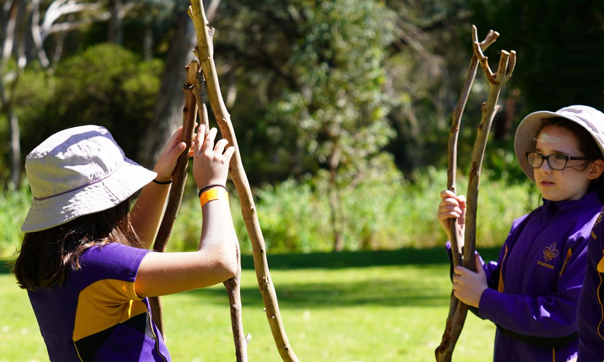 Two young girls play in the grass, each holding a stick and smiling at each other.