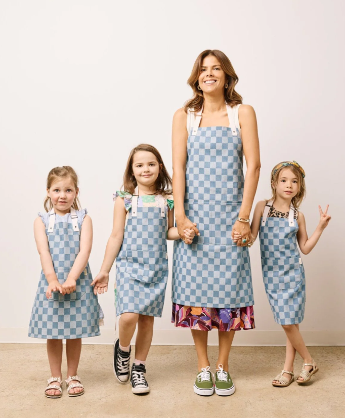 A woman and three children wearing blue aprons smile together, standing in a bright kitchen setting.