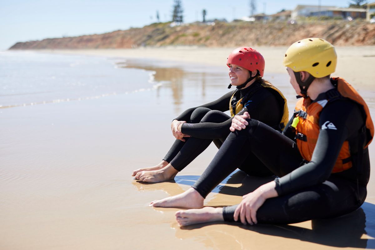  Two people wearing wetsuits relax on the beach, surrounded by sand and the ocean in the background.