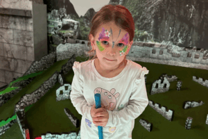 A young girl with colorful face paint smiles while holding a toy in her hand, showcasing her playful spirit.