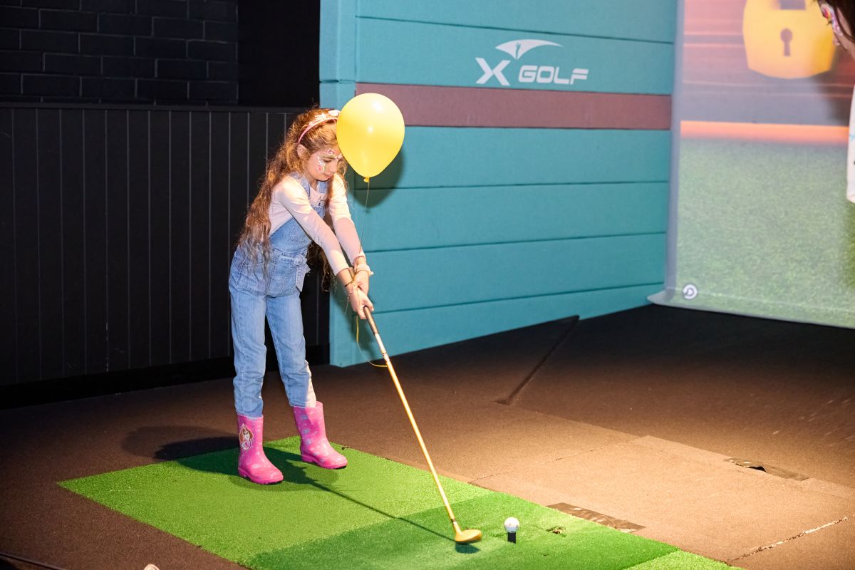 A young girl swings a golf club while playing on a golf simulator, focused on her shot in a virtual environment.
